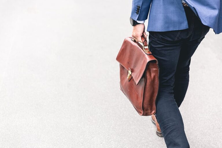 Person walking with a brown leather briefcase, wearing a blue blazer and dark pants, seen from behind on a gray pavement.