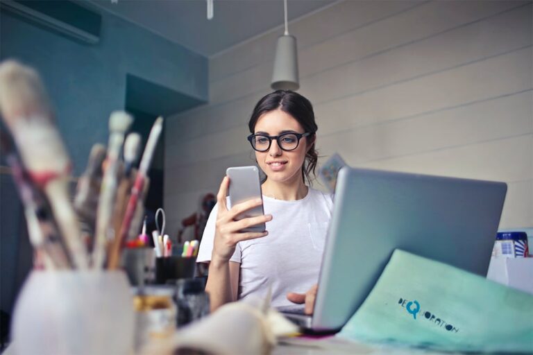 A person wearing glasses sits at a desk with a laptop, holding a smartphone. Art supplies and a cup are on the desk.