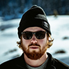 A person with a beard wears sunglasses and a beanie, standing outdoors in a snowy landscape.