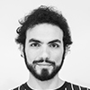 A black and white portrait of a person with curly hair, a beard, and a striped shirt, looking directly at the camera.