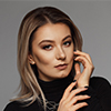 Person with long blonde hair, wearing a black turtleneck sweater, poses against a gray background with one hand touching their face.